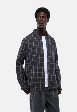 Chemise en flanelle gris à carreaux avec boutons, dotée d'un motif en grille blanche et d'une coupe décontractée. Portée par-dessus un t-shirt blanc uni.