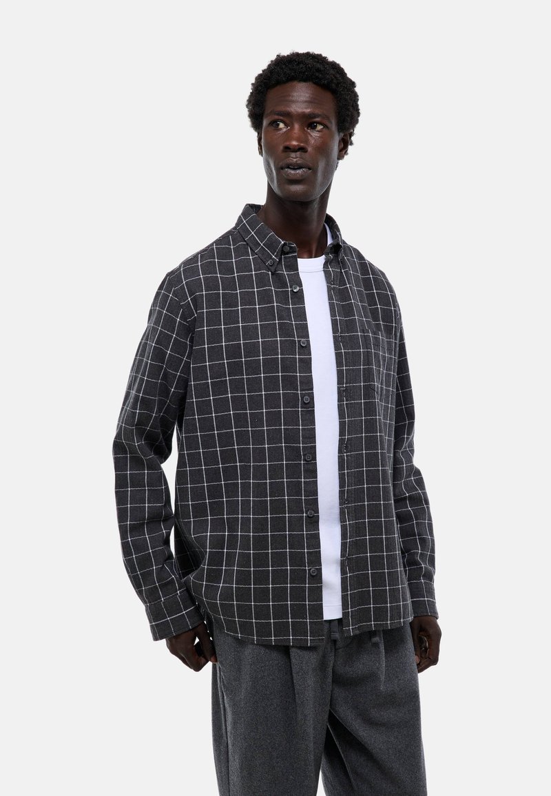 Chemise en flanelle gris à carreaux avec boutons, dotée d'un motif en grille blanche et d'une coupe décontractée. Portée par-dessus un t-shirt blanc uni.