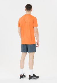 Orange kortärmad skjorta, grå shorts, svarta löparskor med vita strumpor. Skjortan har en slät textur och en avslappnad passform.