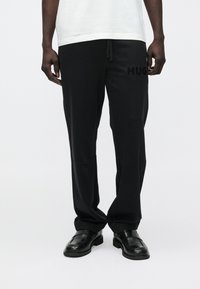 Man die zwarte broek draagt met de tekst "HUG" op het dijbeen, zwarte loafers en een wit overhemd, staand tegen een effen witte achtergrond.