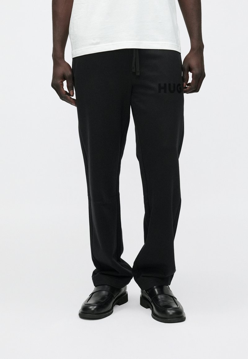 Man die zwarte broek draagt met de tekst "HUG" op het dijbeen, zwarte loafers en een wit overhemd, staand tegen een effen witte achtergrond.