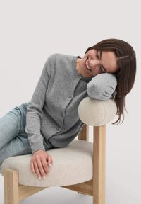 Frau, die den Kopf auf einer gepolsterten Armlehne eines Holzstuhls ruht, mit geschlossenen Augen lächelt, trägt einen grauen Cardigan und blaue Jeans.