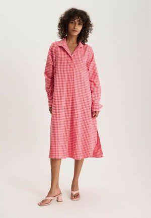 Femme aux cheveux bouclés portant une robe midi à manches longues à carreaux roses et rouges et des sandales à talons roses clairs, debout sur un fond blanc.