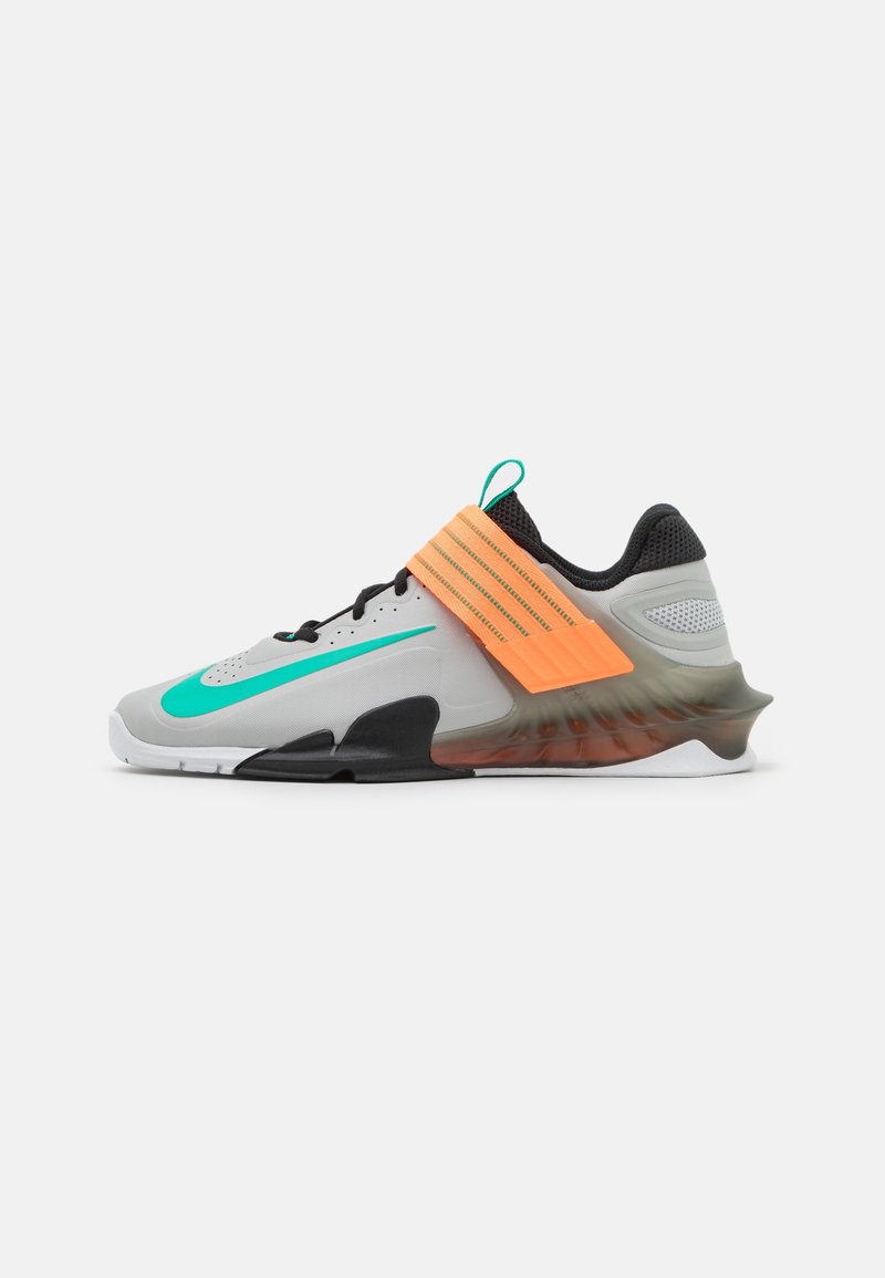 Sportschoen met een grijze bovenkant, zwarte accenten, groen Nike-logo en een oranje band. Kenmerkt zich door een gestructureerde, doorzichtige buitenzool voor grip.