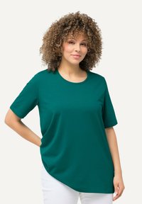 T-shirt corto color teal con scollatura rotonda, vestibilità rilassata e orlo asimmetrico. Realizzato in materiale morbido, indossato sopra pantaloni leggeri.