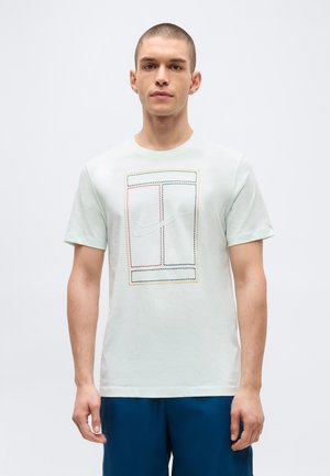 Homme aux cheveux courts portant un t-shirt vert clair avec un motif rectangulaire multicolore cousu et un logo blanc Nike Swoosh, associé à un short bleu.