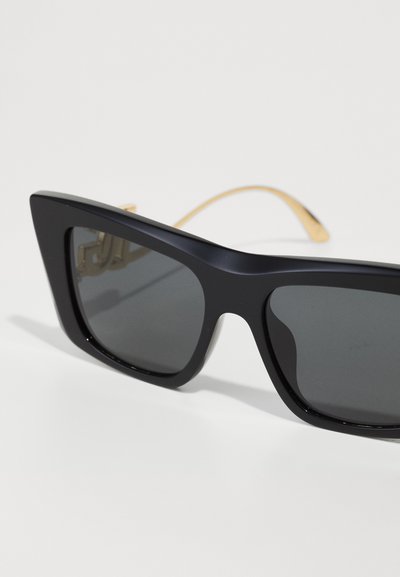 Black oversized sunglasses with a thick frame, dark lenses, and gold-tone metal accents on the temples. Smooth texture, angular design.