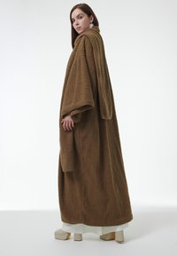 Bata de tela marrón y esponjosa con mangas anchas, dobladillo hasta la rodilla y superficie texturizada. El modelo lleva zapatos de plataforma, mostrando un atuendo en capas.