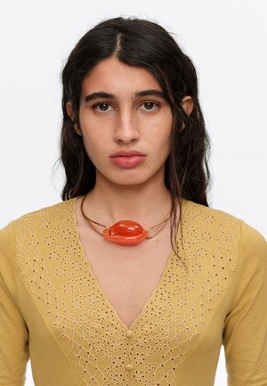 Mujer con cabello largo y oscuro que lleva un vestido amarillo mostaza con bordado floral y un collar rojo de forma abstracta.