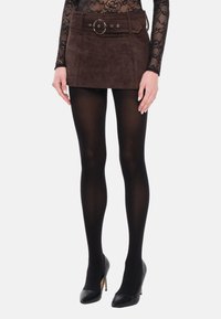 Mini-jupe en daim marron avec une large ceinture dotée d'une boucle circulaire, associée à des collants opaques noirs et des chaussures à talons hauts noires.