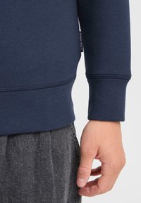 Mano derecha al lado del cuerpo, con sudadera azul marino que tiene la etiqueta "Casual Friday" y pantalones grises con textura.