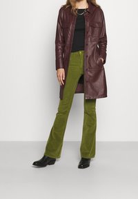 Manteau long en cuir bordeaux avec deux poches poitrine, associé à un pantalon patte d'eph en velours côtelé vert et des bottines noires à talons.