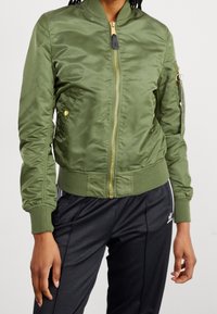 Blouson aviateur vert olive en tissu lisse et léger, avec une fermeture éclair dorée, des poignets côtelés et une poche utilitaire sur la manche.