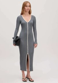 Graues, geripptes Strickkleid mit langen Ärmeln, V-Ausschnitt, Vorderknöpfen, seitlichem Schlitz und kontrastierendem weißen Besatz, kombiniert mit einer schwarzen Clutch.