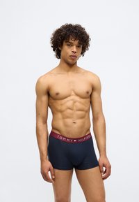 Mladic s kovrčavom kosom nosi mornarskoplave boxer gaće Tommy Hilfiger s bordo pojasom na jednobojnoj bijeloj pozadini.