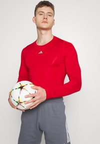 Red long-sleeve athletic shirt with a crew neck and Adidas logo, paired with gray athletic pants. The model holds a patterned soccer ball.