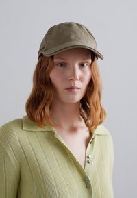 Cappello di cotone verde con visiera curva. Indossato con una camicia colletto di un verde chiaro, dalla texture, con bottoni e sottili righe verticali.