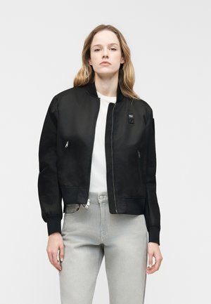 Femme aux longs cheveux blonds portant une veste bomber noire sur une chemise blanche et un jean gris clair, sur un fond clair uni.
