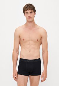 Schwarze Baumwoll-Boxershorts mit glatter Textur, ausgestattet mit einem schwarzen Logo-Gummibund und körpernahem Schnitt. Das Model posiert mit freiem Oberkörper.