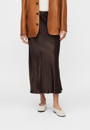 Person iført en brun læderjakke, mørkebrun satin midi-nederdel og lysebeige flade sko, stående mod en ensfarvet baggrund.