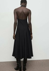 Vestido negro de cuello halter con cuerpo ajustado y falda acampanada, que presenta un diseño de espalda baja y una textura de tela suave. Llevado con botas hasta la rodilla.