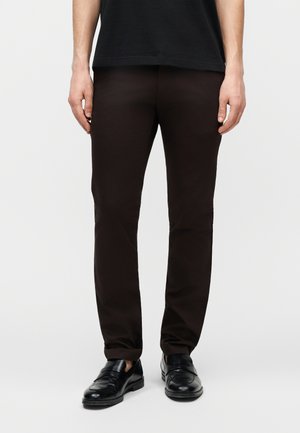 Uomo che indossa pantaloni neri aderenti e mocassini in pelle nera, in piedi con le braccia rilassate lungo i lati contro uno sfondo semplice.