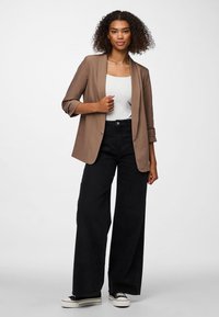 Mujer con cabello rizado que lleva una chaqueta marrón sobre una blusa blanca, pantalones negros de pierna ancha y zapatillas de plataforma en blanco y negro, parada con confianza.