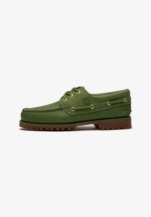 Chaussure bateau en cuir vert avec semelle à crampons marron, lacets passant par des œillets dorés, et logo arbre embossé sur le côté.