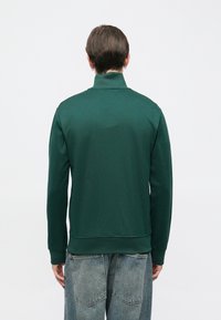 Άνδρας με κοντά καστανά μαλλιά, φοράει σκούρα πράσινη μπλούζα με ψηλό γιακά και ξεθωριασμένο γαλάζιο τζιν, εμφανίζεται από πίσω με φόντο λευκό.