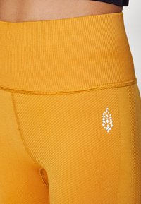 Primer plano de leggings seamless de cintura alta en color mostaza con tejido texturizado y pequeño logo en forma de hoja blanca en la cadera.