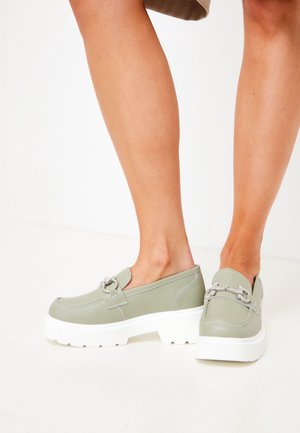 Jambes portant des mocassins vert clair à enfiler avec des semelles blanches épaisses et un détail de chaîne argentée, debout sur un sol blanc.