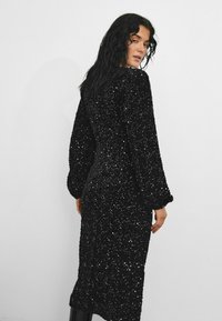Rochie neagră cu mâneci lungi, cu o siluetă ajustată, dintr-un material texturat decorat cu paiete, de lungime medie la nivelul gambei.