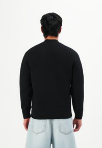 Homme aux cheveux bruns portant un pull noir à manches longues uni et un short en denim bleu clair, se tenant contre un fond blanc.