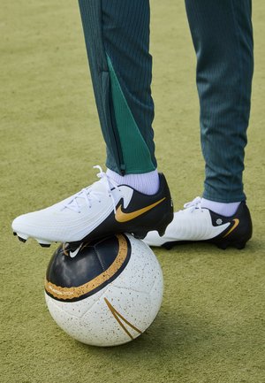 Nike fodboldstøvler, hvide og sorte med en gylden swoosh, hvilende på en sort og hvid fodbold med sprøjtede accenter. Grønne sportsbukser synlige.