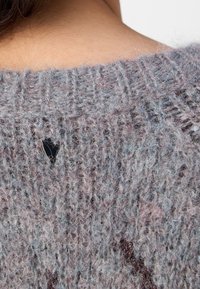 Detailní záběr na krk a rameno osoby oblečené v fuzzy šedém pleteném svetr, na kterém je připevněná malá černá srdíčková brož.