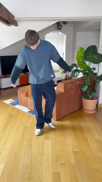 A person wears a blue oversized sweatshirt with black sleeves and navy corduroy pants, standing on a wooden floor near a green potted plant.