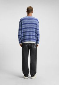 Letvægstriktrøje i blå med horisontale striber, parret med løse sorte jeans og sko med hvid sål, set fra ryggen.