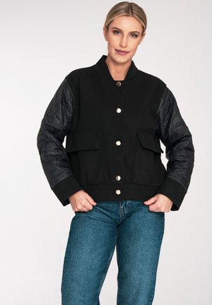 Schwarze Bomberjacke mit gepolsterten schwarzen Ärmeln, Rundhalsausschnitt, goldener Knopfverschluss und zwei Fronttaschen. Weicher Stoff mit kontrastierender Textur.