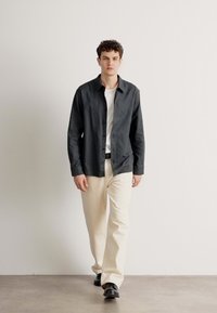 Camicia scura sbottonata sopra una maglietta bianca, pantaloni beige chiaro e scarpe nere; il modello cammina su uno sfondo neutro.