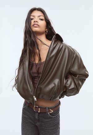 Braune Fake-Leder-Übergangsjacke in Übergröße mit einem Reißverschluss, breitem Kragen und weit geschnittenen Ärmeln, getragen über einem gerippten braunen Oberteil und dunklen Jeans.