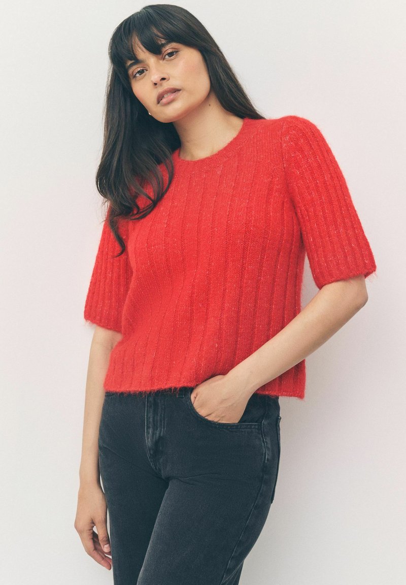 Röd ribbad stickad tröja med korta ärmar, med en mjuk textur och en cropped design. Bärs med svarta jeans, händerna i fickorna.
