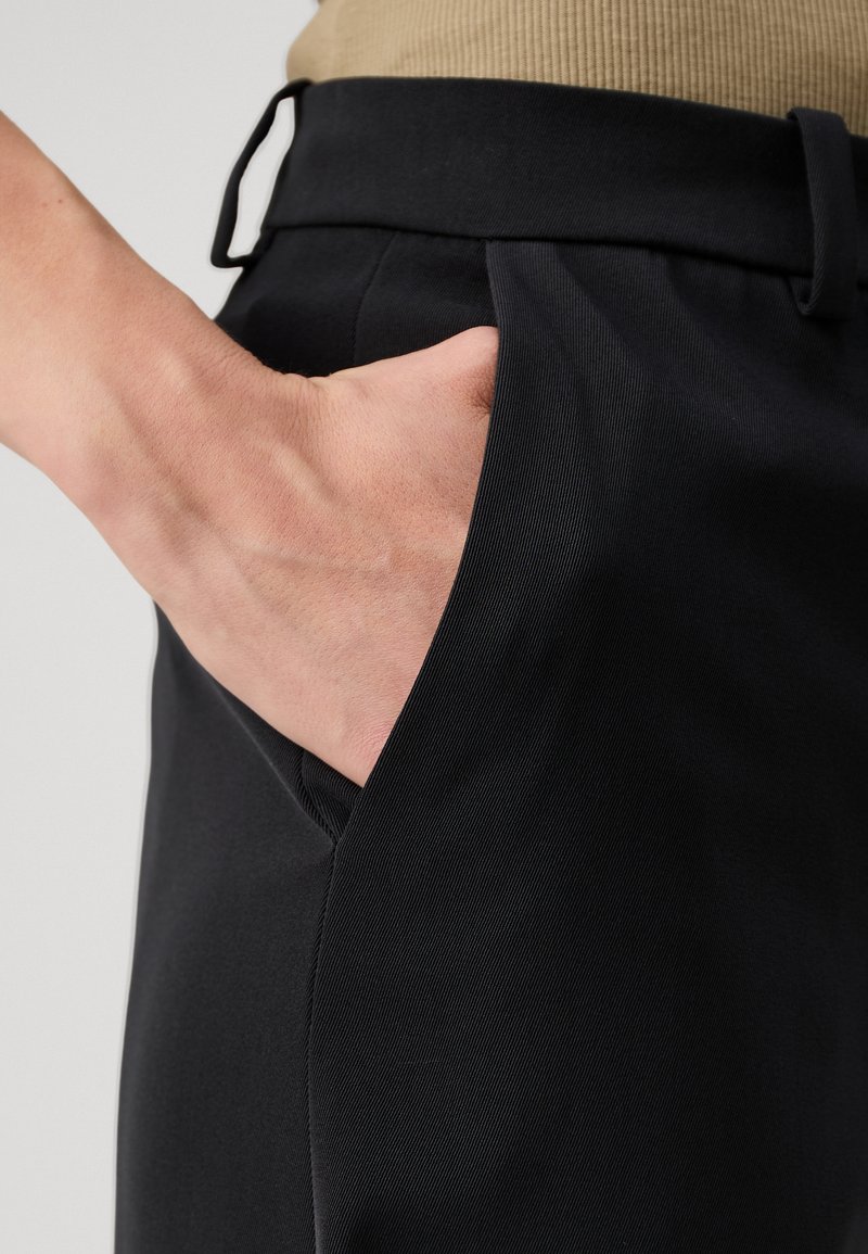 Zwarte op maat gemaakte broek met een gladde textuur, voorzien van een zijzak en een nauwsluitende tailleband. Close-up van een hand die in de zak is geplaatst.