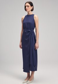 Navy sleeveless dress with a gathered waist and side knot detail, made from lightweight fabric. Model wearing pale pointed-toe flats.