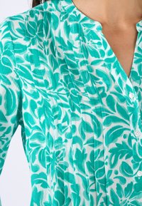 Korte mouwen top in een turkooizen bloemenmotief op een witte achtergrond, met een knoopsluiting aan de voorkant, plooien en een kraag.