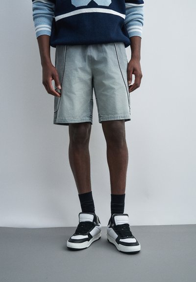 Homme portant un short gris clair avec des poches latérales, des chaussettes noires, des baskets noir et blanc, et un pull bleu marine à rayures bleu clair.
