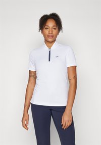 White polo shirt with short sleeves, half zip neck, and CK logo; paired with navy blue pants; smooth, lightweight fabric.