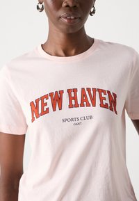 Pats šviesiai rožinės spalvos medvilnės marškinėliai su ryškiu raudonu užrašu "NEW HAVEN", mažesniu užrašu "SPORTS CLUB" ir "GANT" žemiau, su klasikiniu apvaliu kaklu ir trumpomis rankovėmis.