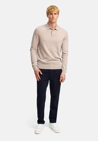 Hellbeige langärmliges Polohemd mit einem Kragen, kombiniert mit navyblauen Hosen und weißen Sneakern, vor einem schlichten Hintergrund stehend.
