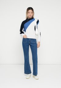 Strikkegenser med marineblå, hvite og lyseblå geometriske mønstre; sammen med lyseblå utsvingte jeans og hvite sneakers.