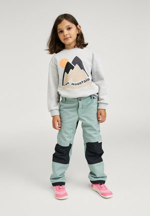 Young girl standing wearing grey mountain-print sweatshirt, light blue pants with black patches, and pink sneakers against plain background.